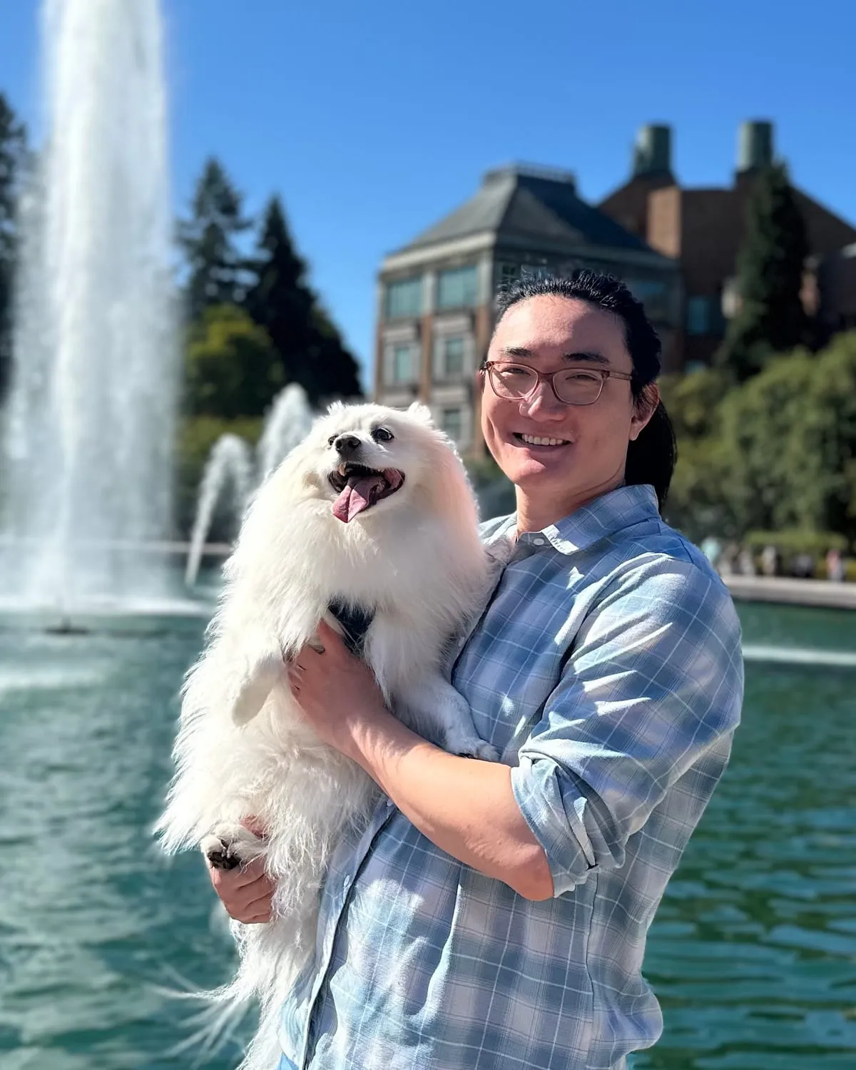 Sean, in glasses and a plaid shirt, holding Dotes, a fluffy white dog, in front of a fountain on a sunny day